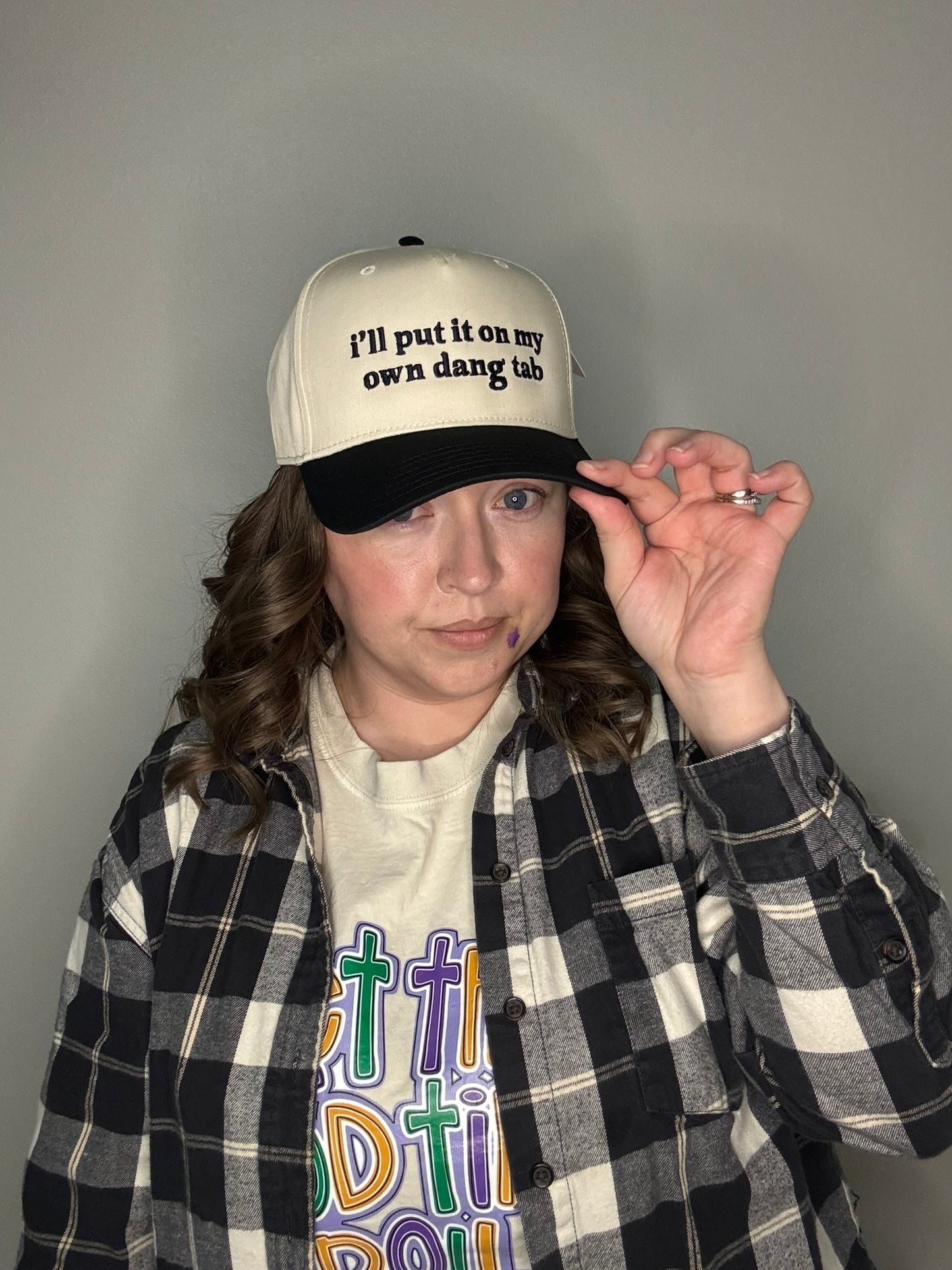 Person wearing a white cap with "I'll put it on my own dang tab" in black text, plaid jacket, and colorful shirt against a gray background
