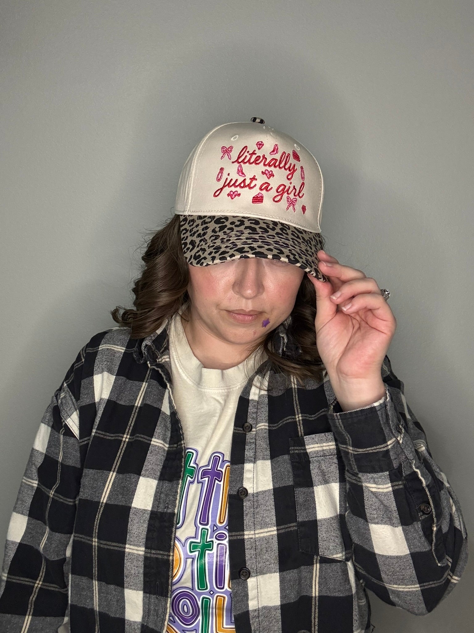 Person wearing a hat with "literally just a girl" text in red font, plaid jacket, and colorful shirt against a plain background