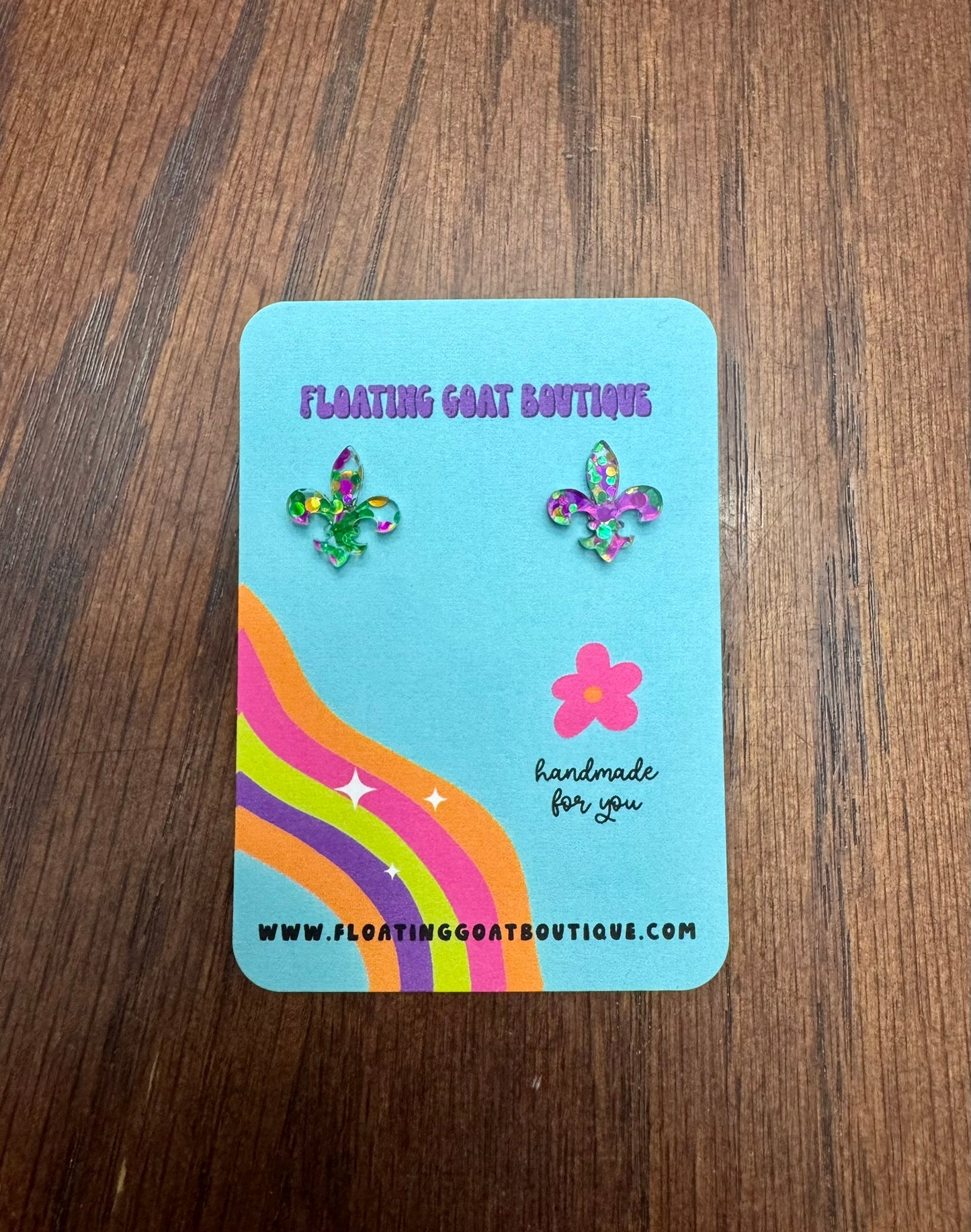 Pair of Fleur de Lis Mardi Gras Confetti earrings on a blue card with rainbow design on a wooden surface.