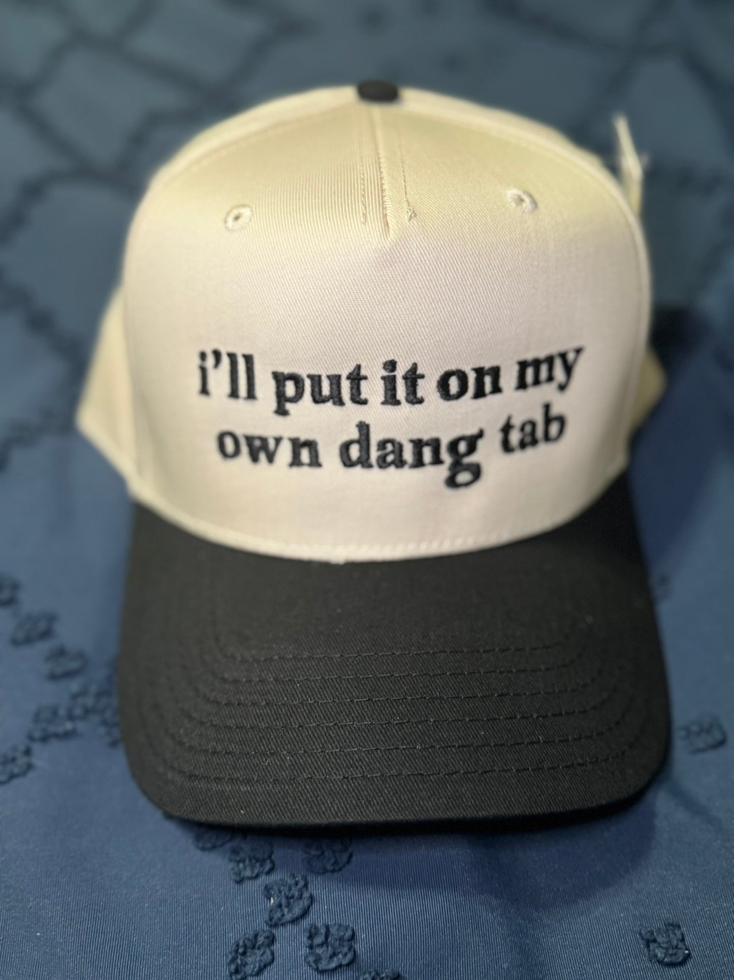 Beige and black cap with "I'll put it on my own dang tab" in black text on a textured blue background