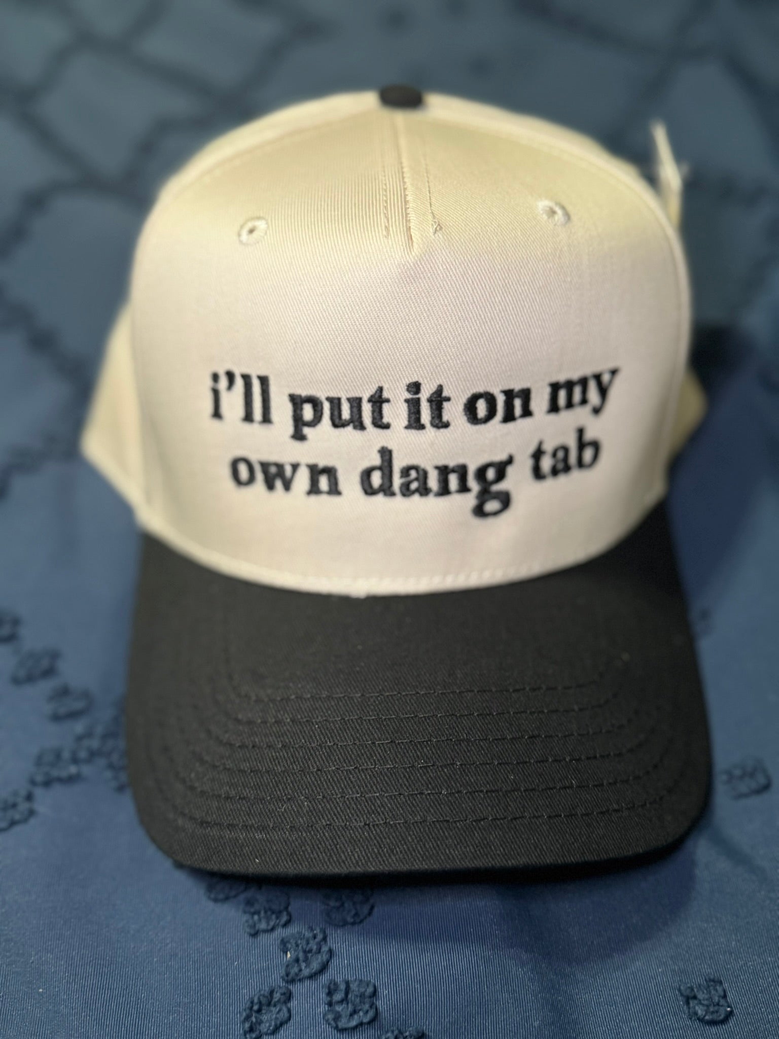Beige and black cap with "I'll put it on my own dang tab" in black text on a textured blue background