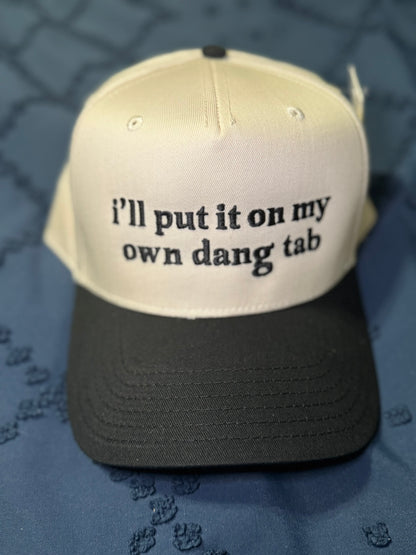 Beige and black cap with "I'll put it on my own dang tab" in black text on a textured blue background