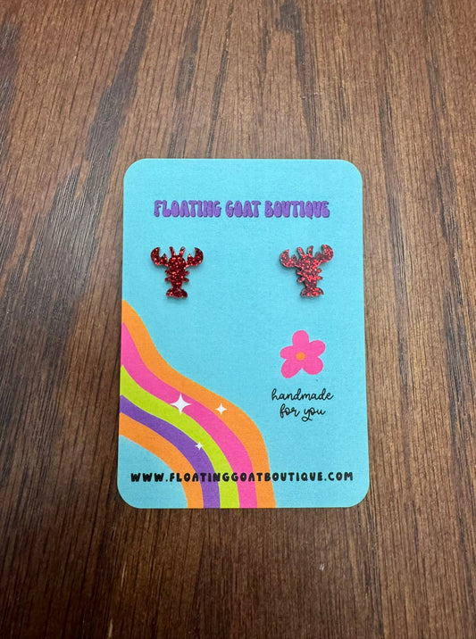 Pair of red crawfish earrings on a blue card with colorful design, displayed on a wooden surface.
