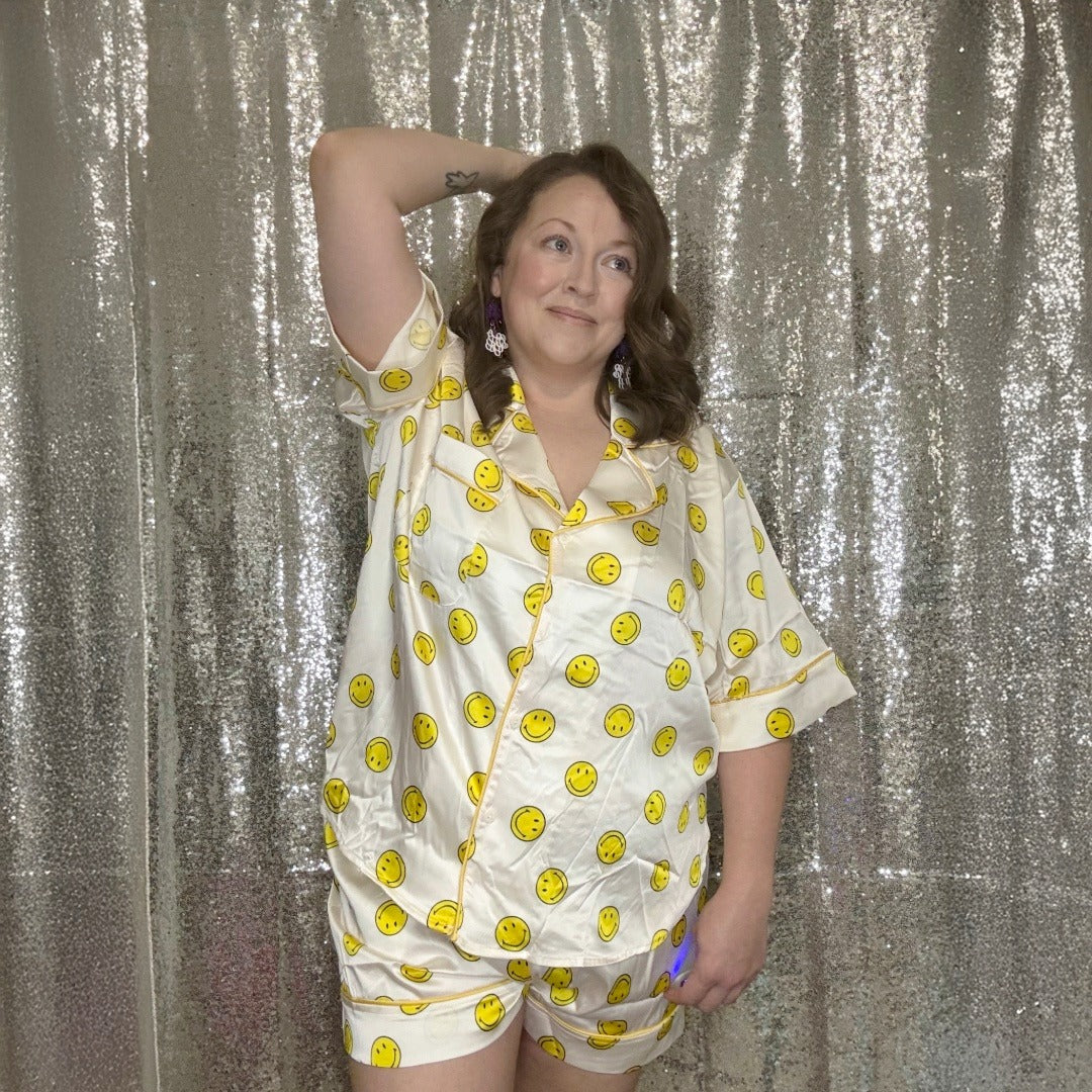 Person wearing white satin pajamas with yellow smiley face patterns against a silver glittery curtain.