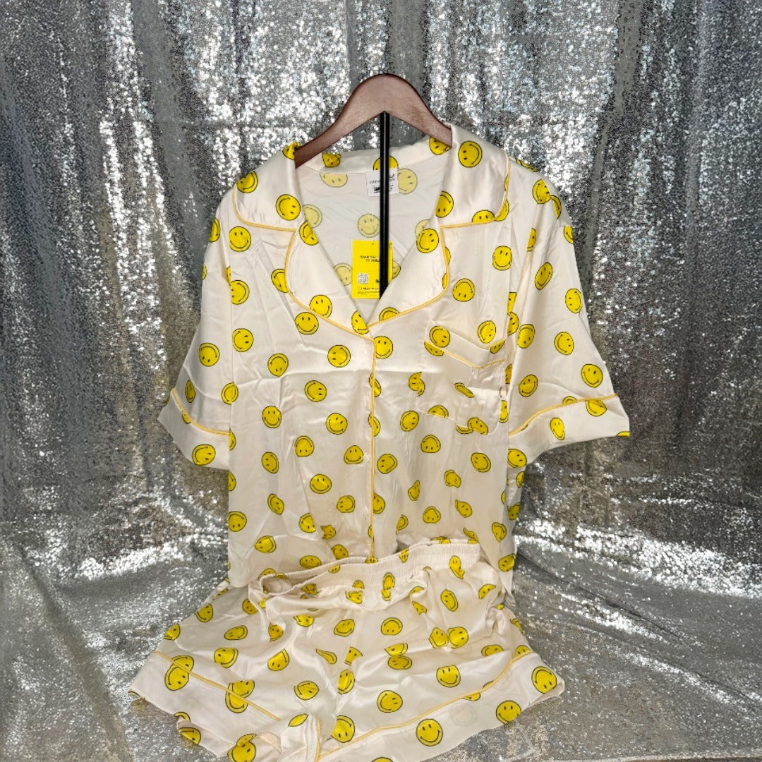 White satin short pajamas with yellow smiley face patterns on a hanger against a silver curtain background